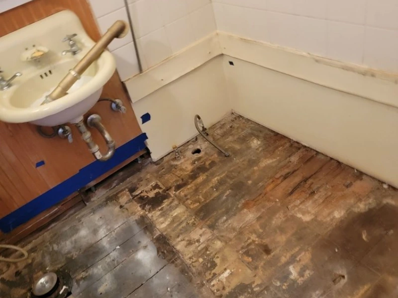 Bathroom corner with an old sink and a worn, partially stripped wooden floor.