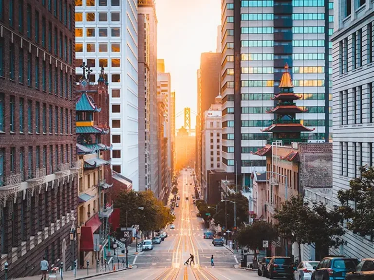 Sunset view down a city street lined with tall buildings and a distant bridge.